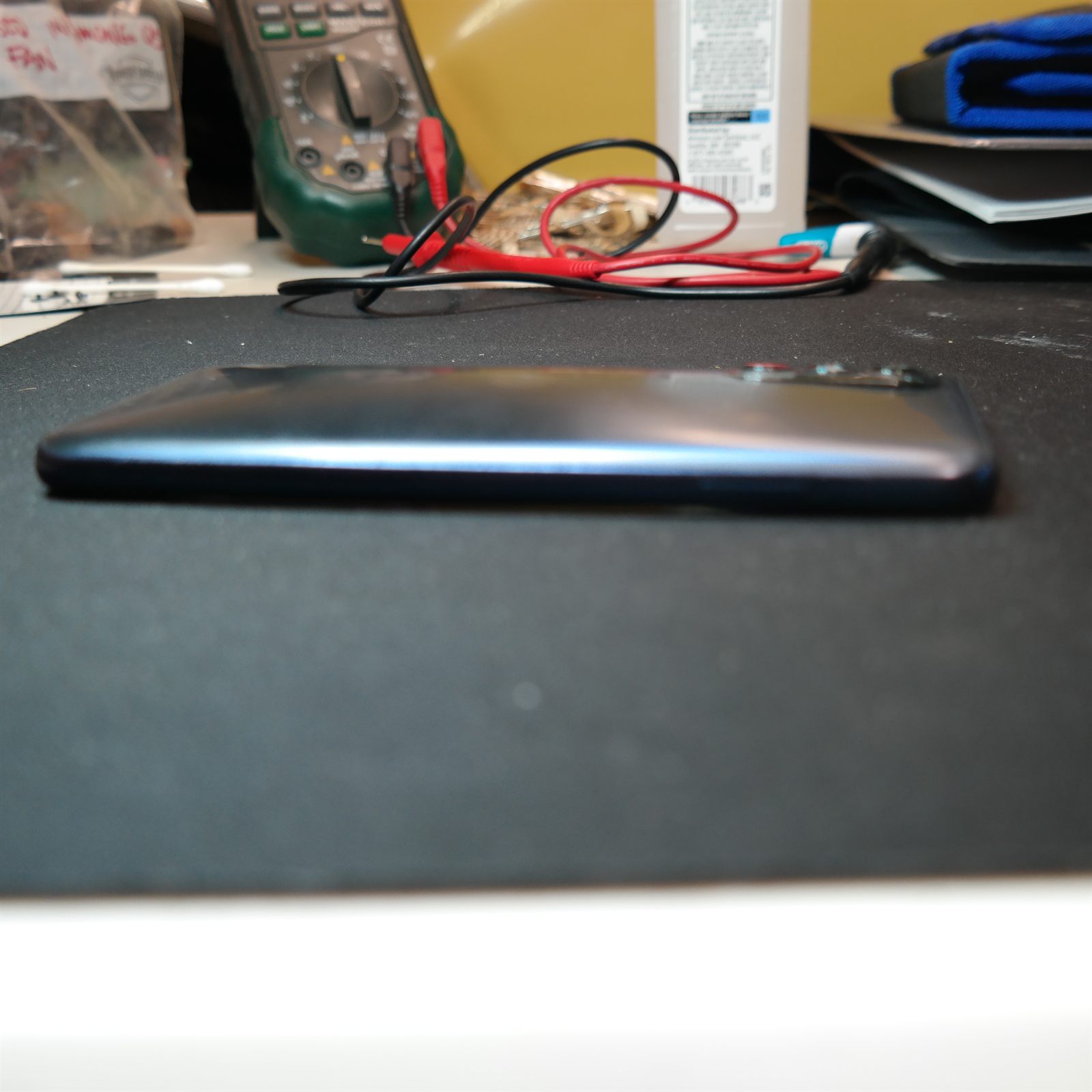 Side profile of the closed Moto G 5G (2022) lying on a black repair mat as it arrived, photographed along the long edge where the rear cover was lifting away from the frame. A multimeter and test leads sit on the bench behind it.