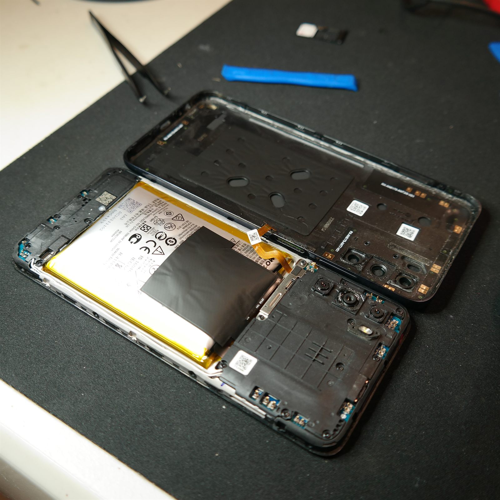 The Moto G 5G (2022) opened like a book on a black repair mat. The rear cover lies to the right, still joined to the phone by a short orange power-button flex cable. The battery and internal cover are visible in the phone body, with tweezers and a blue plastic opening tool on the bench above.